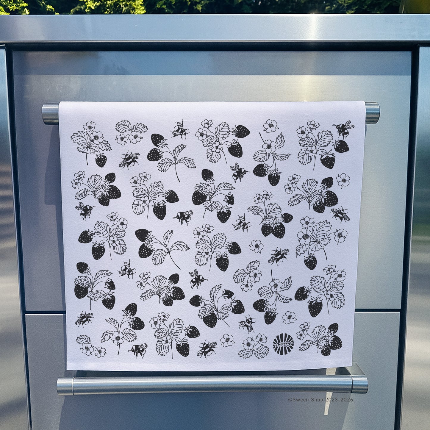White towel with black floral strawberry and bumble bee pattern hanging on a stainless steel appliance