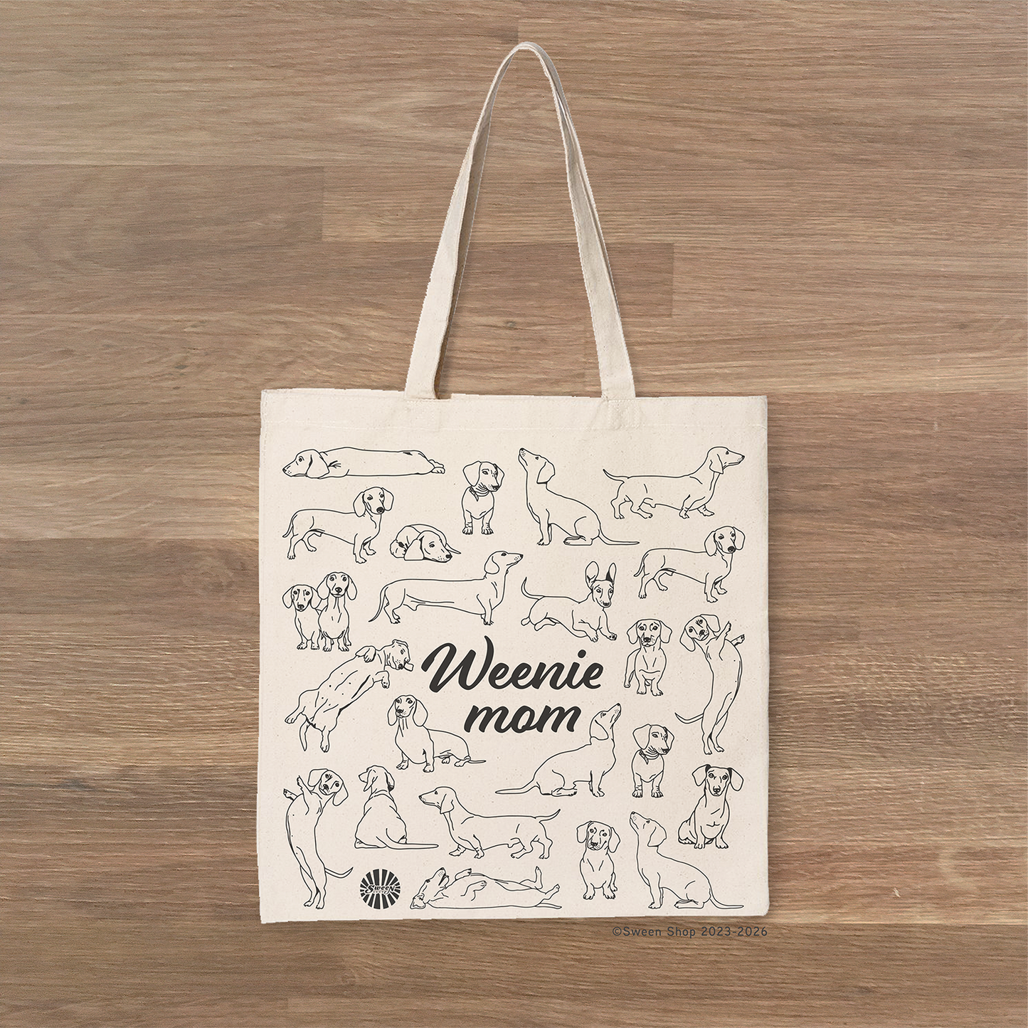 Tote bag with 'Weenie mom' text and Dachshund dog illustrations on a wooden surface