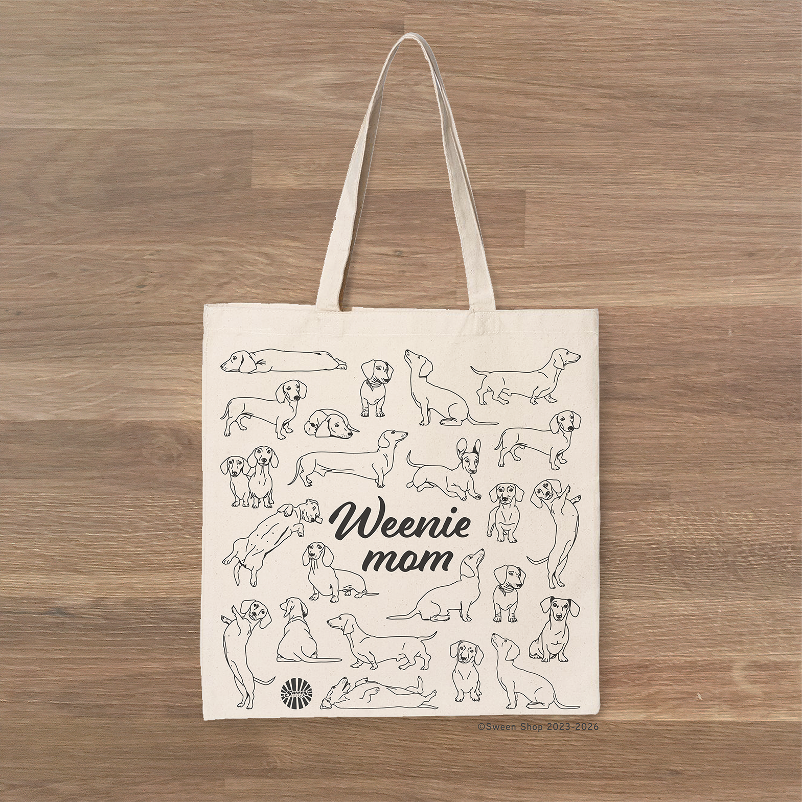 Tote bag with 'Weenie mom' text and Dachshund dog illustrations on a wooden surface