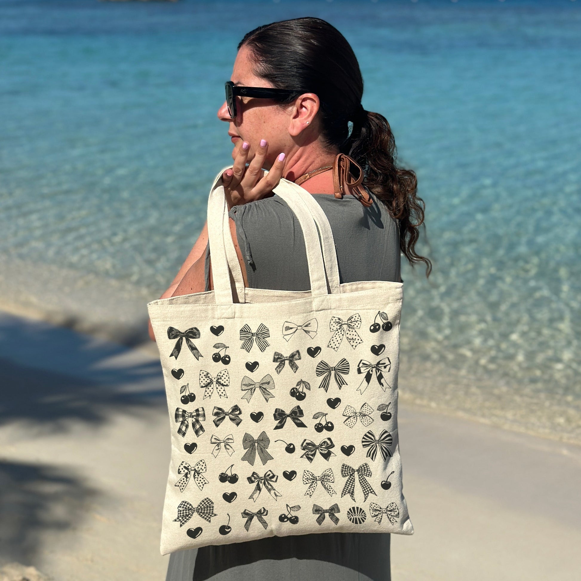 Woman holding a natural cotton canvas tote bag with heart bow cherry hand drawn printed pattern on a beach