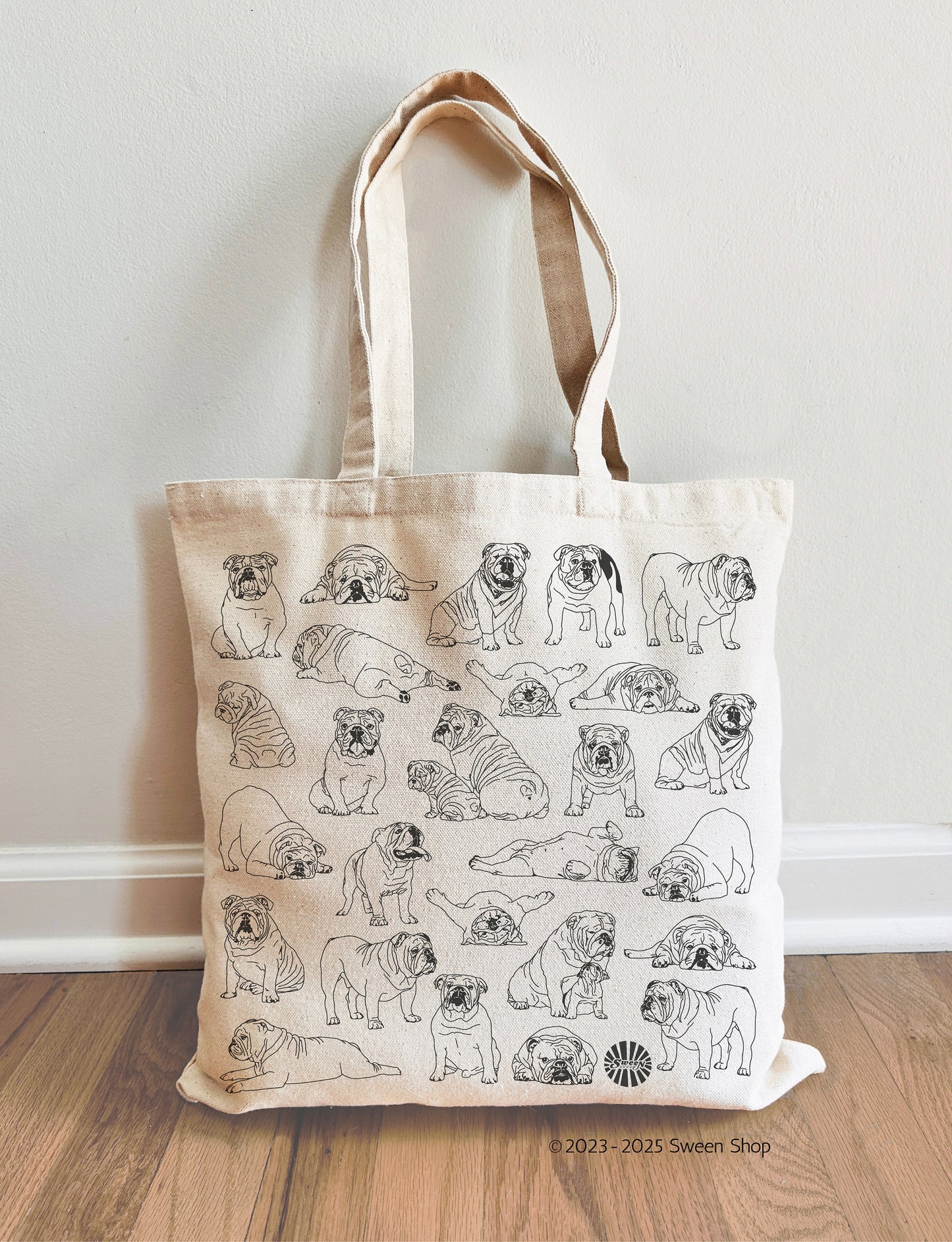 Tote bag with bulldog pet dog illustrations on a wooden floor against a white wall.