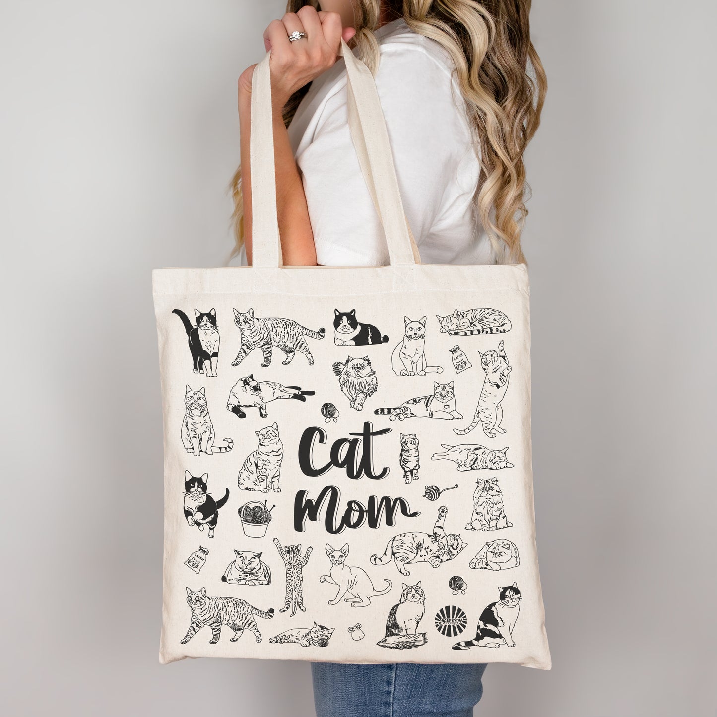 Tote bag with black pet cat illustrations and 'Cat Mom' text held by a person against a plain background