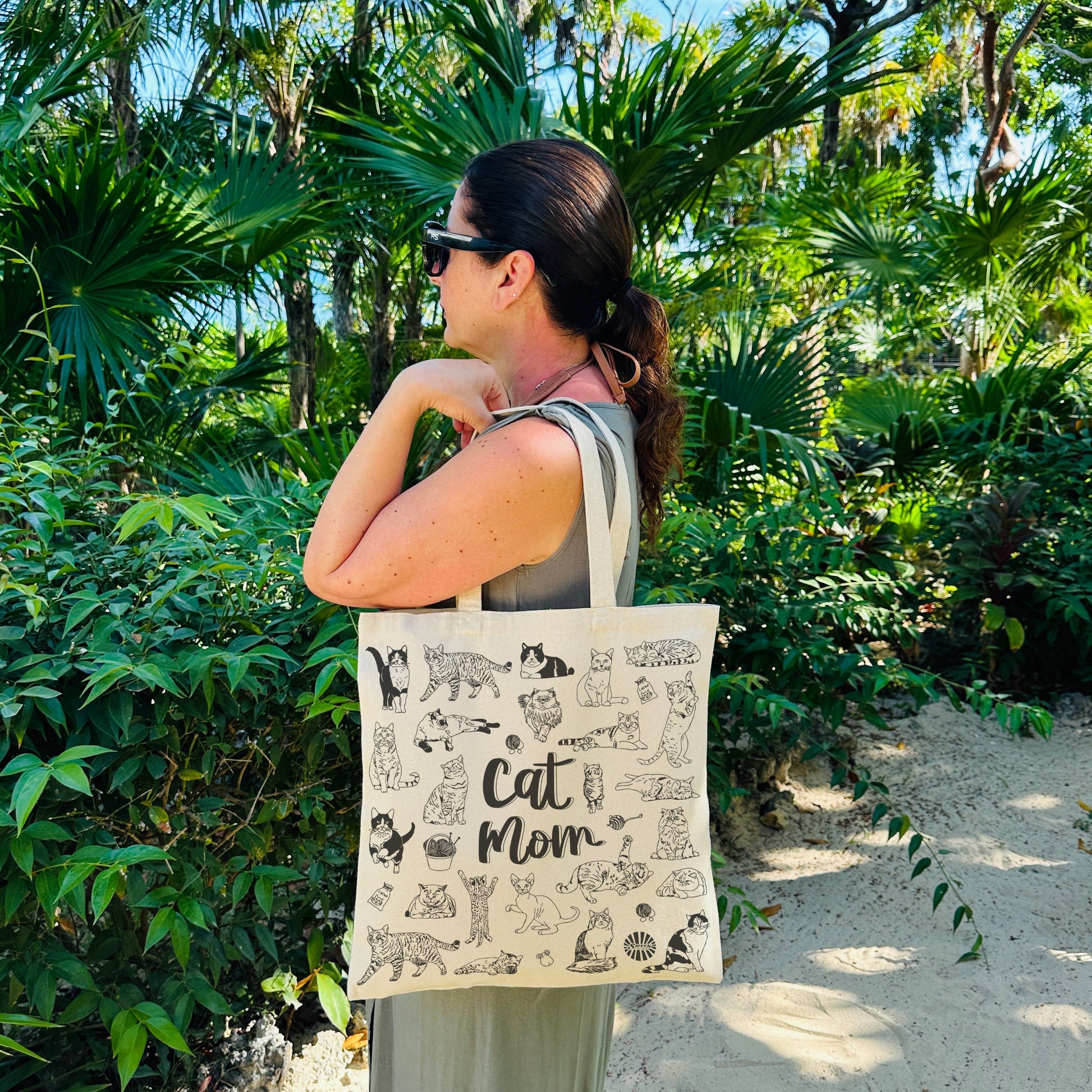 Woman holding a tote bag with 'Cat Mom' design in a tropical setting