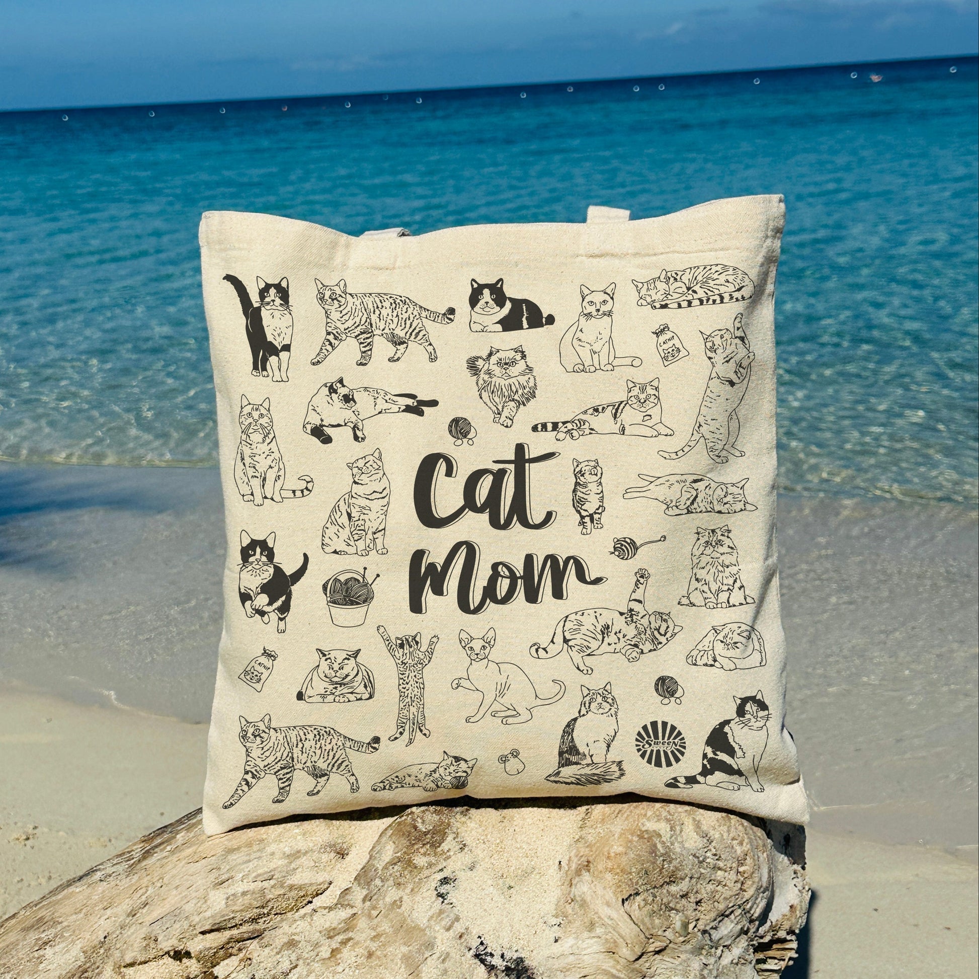 tote bag with 'Cat Mom' text and cat illustrations on a rock by the ocean