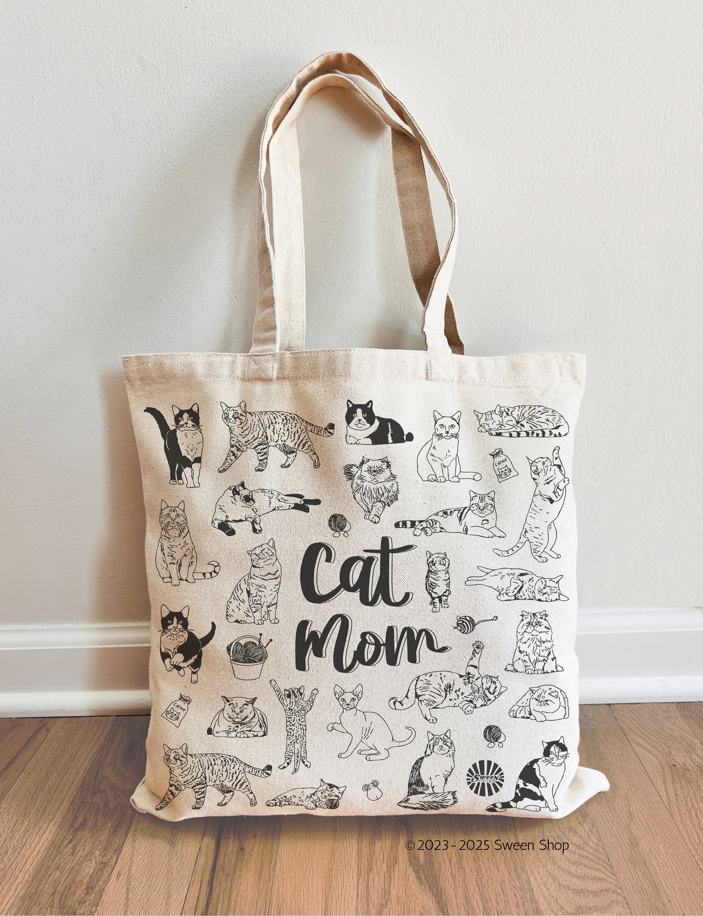Tote bag with cat illustrations and 'Cat Mom' text on a wooden floor.