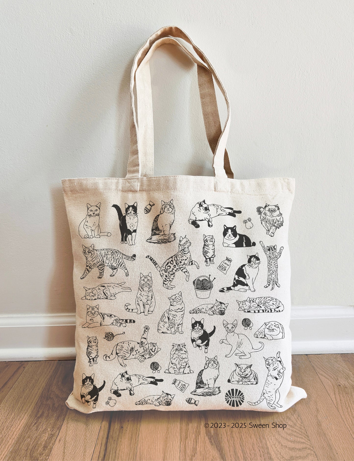 Beige cotton canvas tote bag with black pet cat illustrations on a wooden floor against a white wall.