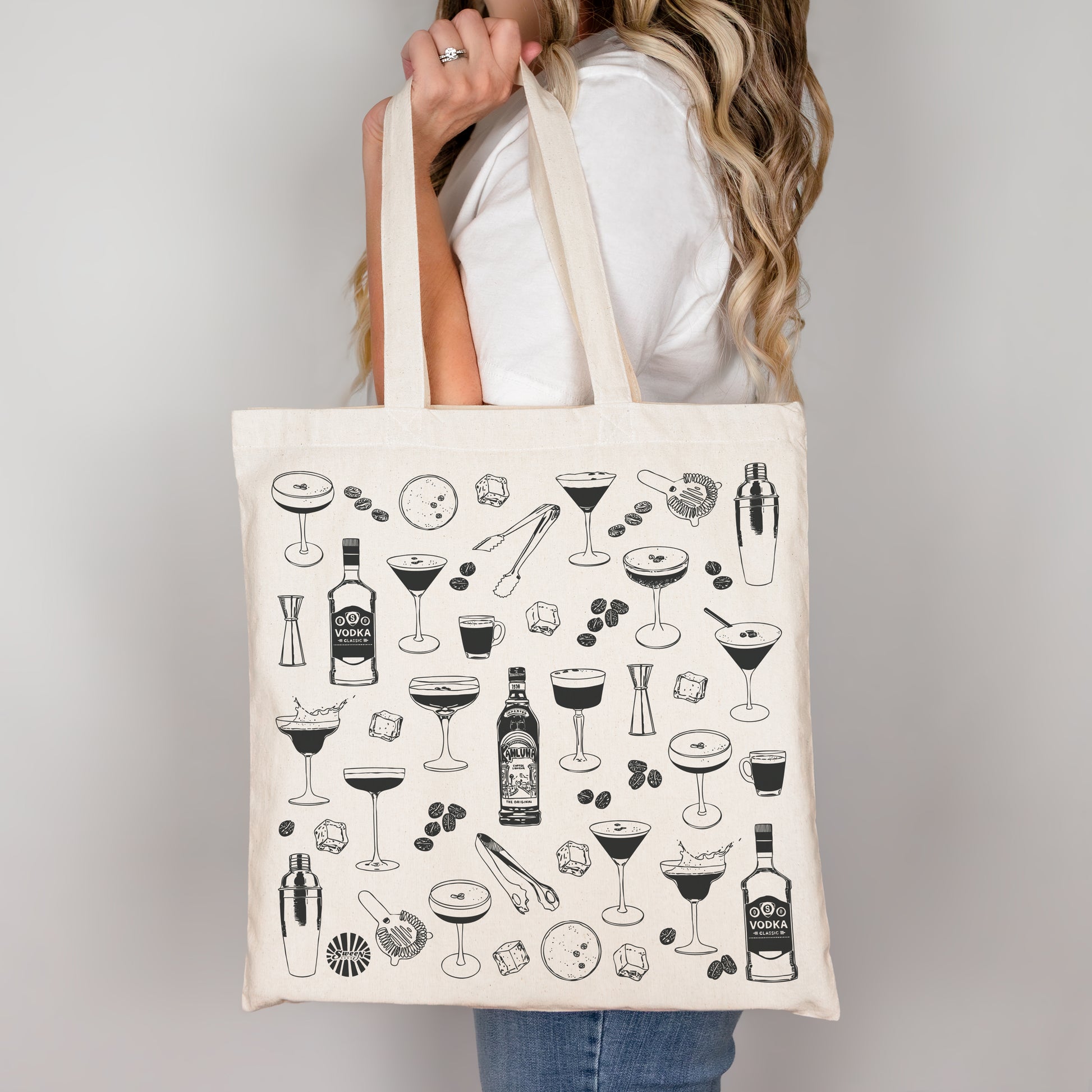 Tote bag with black espresso martini cocktail illustrations held by a person against a plain background