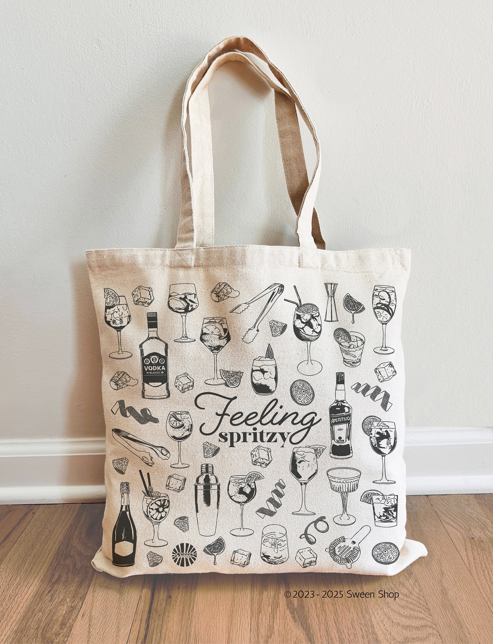 Tote bag with aperol spritz cocktail drink illustrations and 'Feeling spritzy' text on a wooden floor.