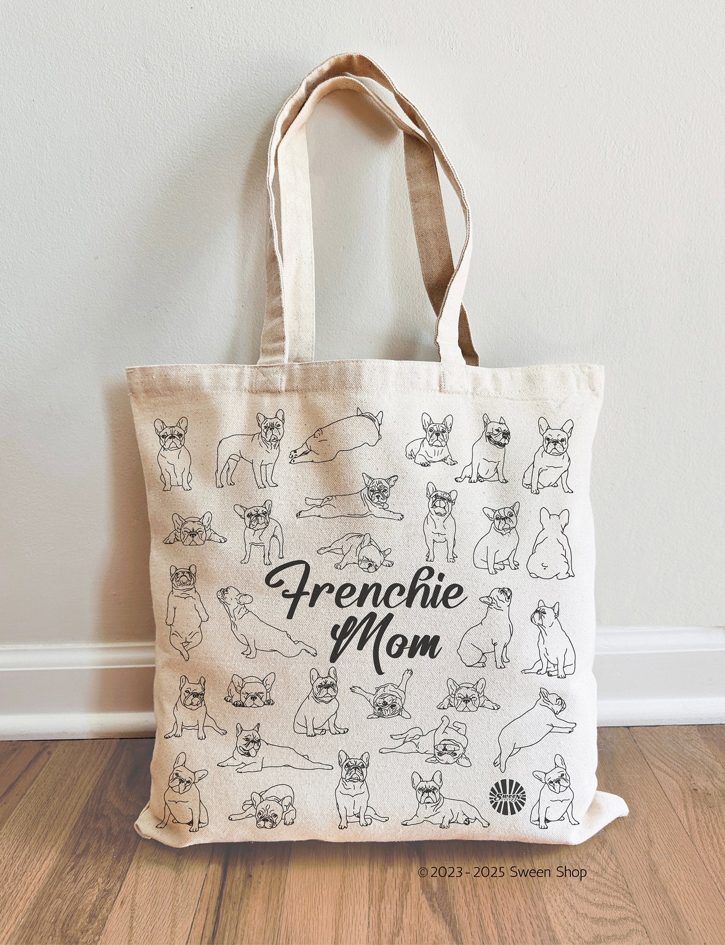 Tote bag with 'Frenchie Mom' text and french bulldog pet dog illustrations on a wooden floor.