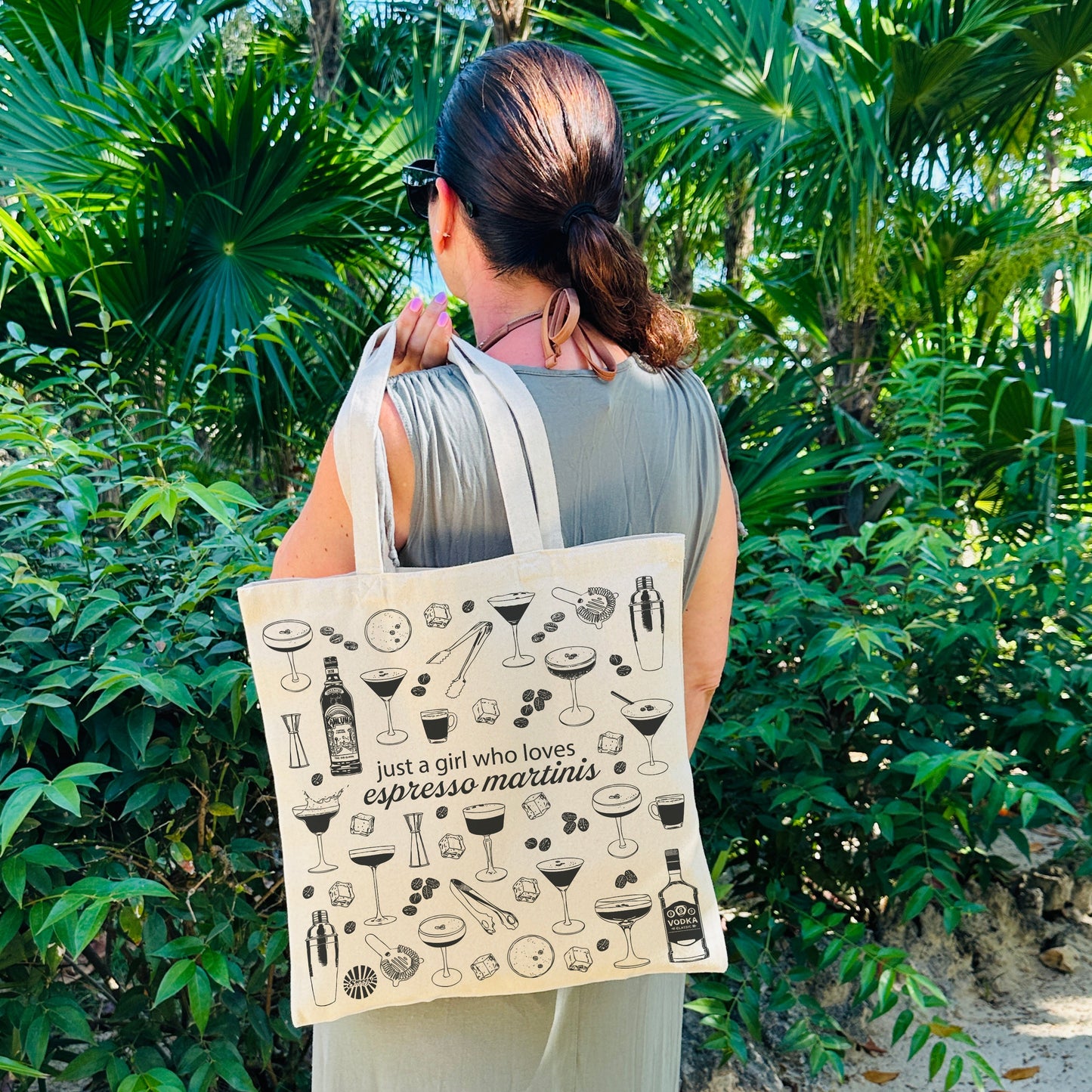 Woman holding a tote bag with espresso martini cocktail drink illustrations and text in a tropical setting