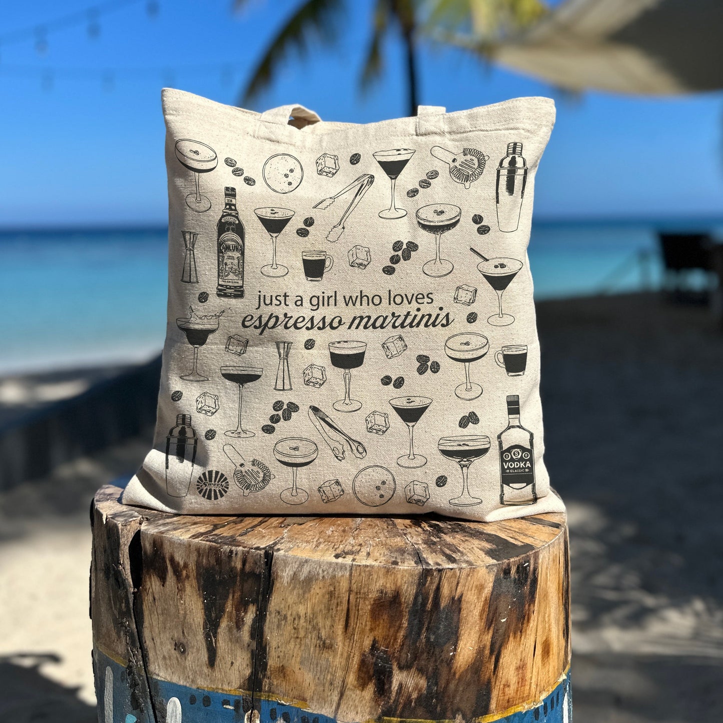 beach tote bag with espresso martini cocktail-themed design on a wooden post by a beach with palm trees.