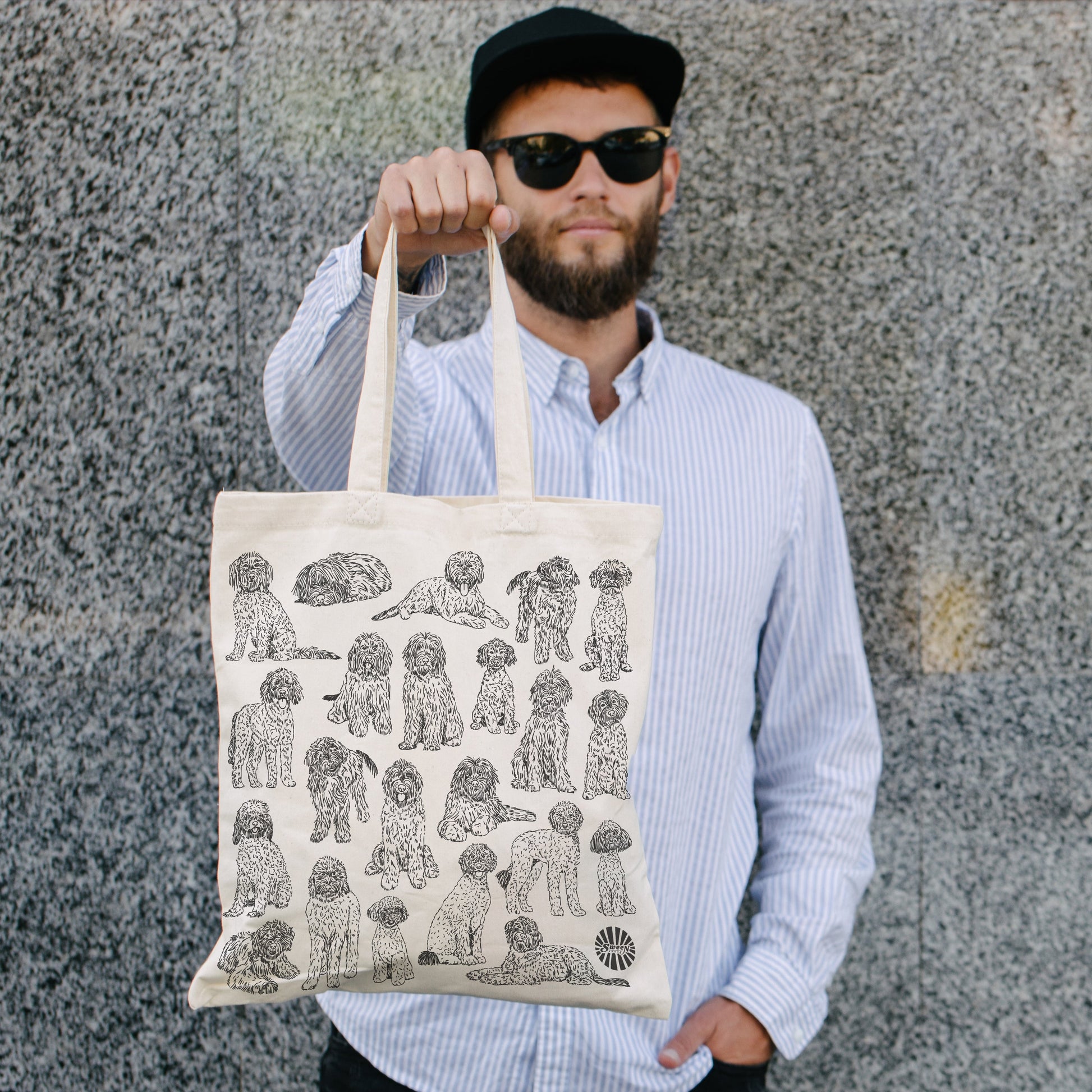 Man holding a tote bag with pet golden doodle dog illustrations against a stone wall.