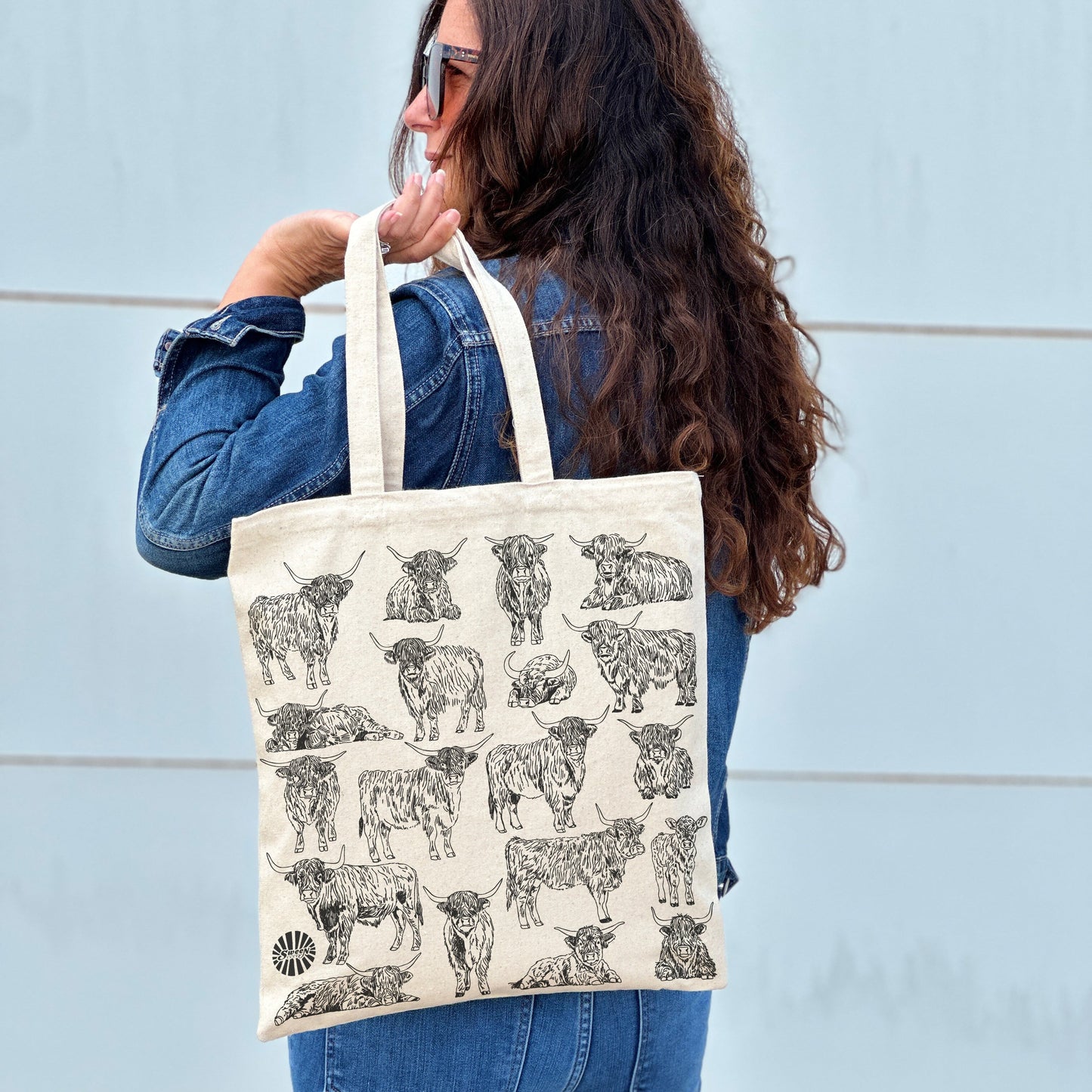 Person holding a tote bag with highland cow pattern against a light blue wall