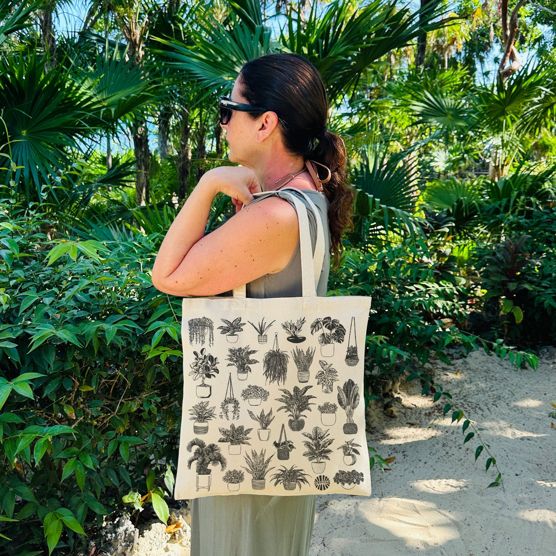 Woman walking through a tropical garden with a tote bag featuring plant designs.