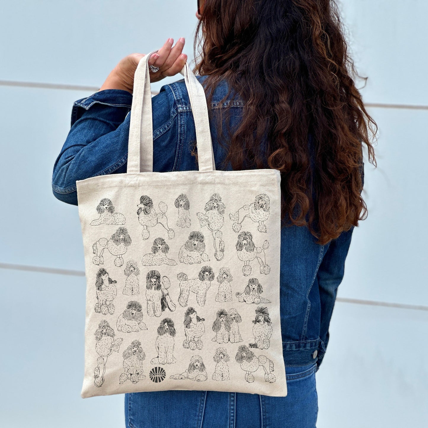 Person holding a tote bag with poodle pet dog illustrations against a light blue wall.