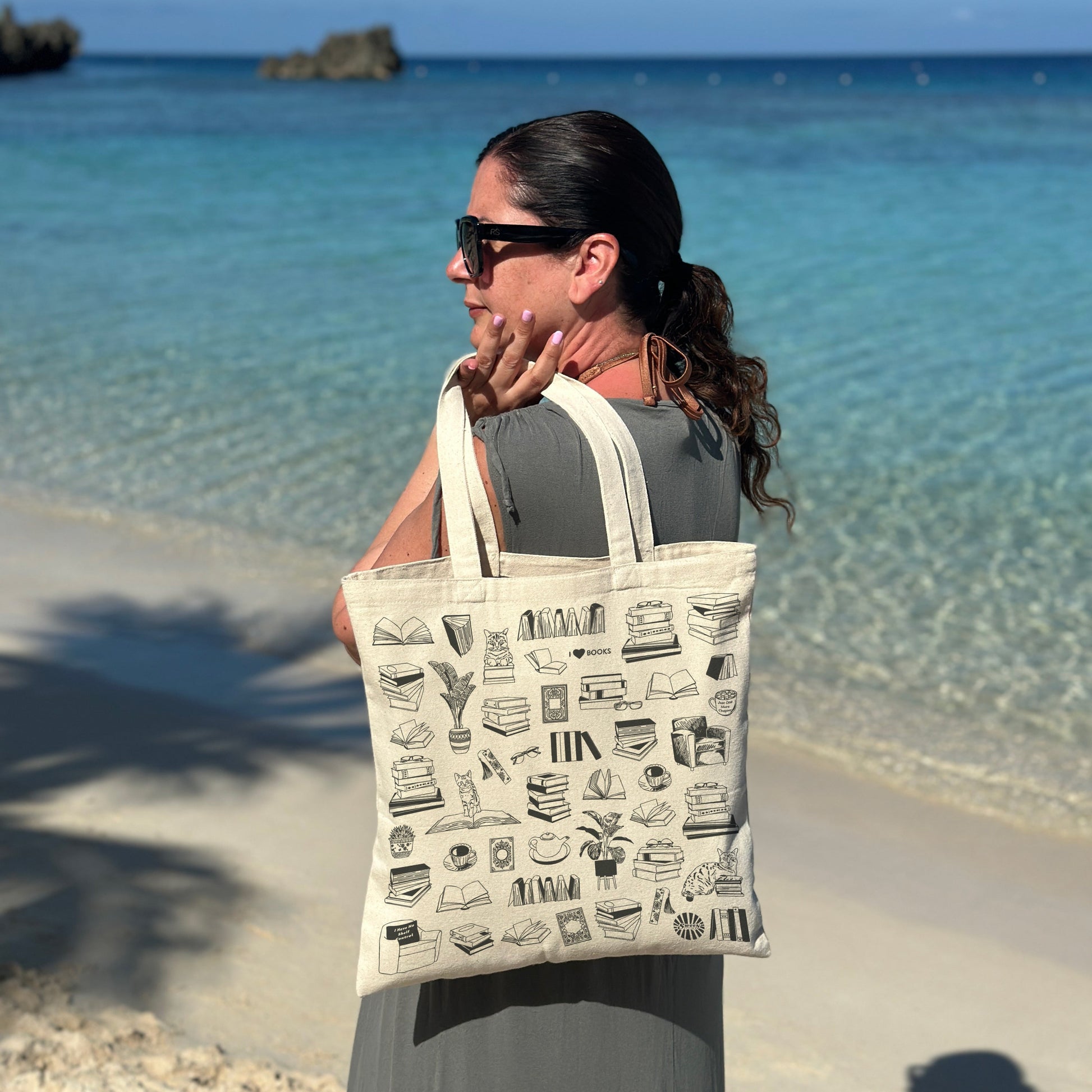 Woman holding a beach tote bag with book reading themed designs by the ocean.