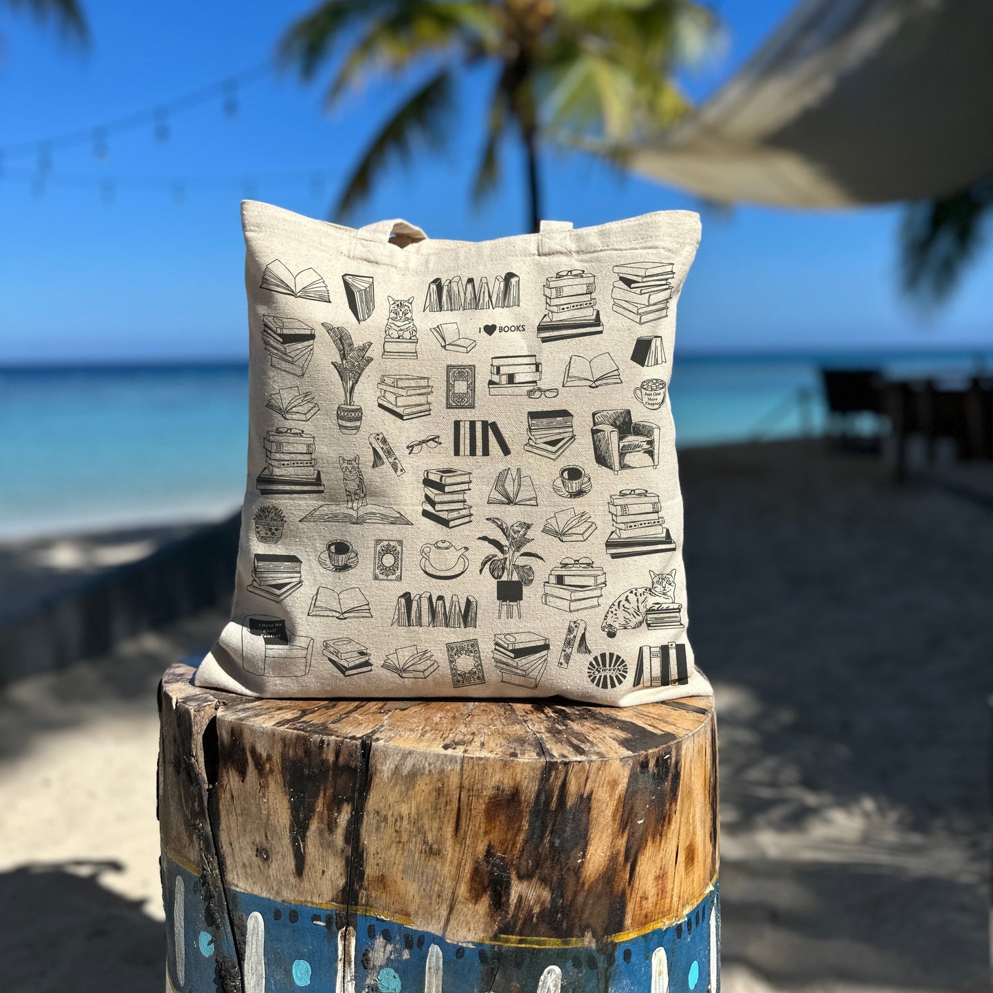 cotton canvas tote bag with black printed reading book theme on a wooden post by a beach with palm trees.
