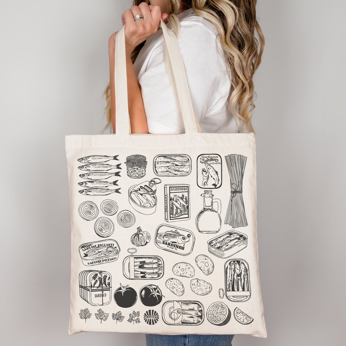 Person holding a tote bag with black sardine pasta canned fish tomato illustrations on a plain background
