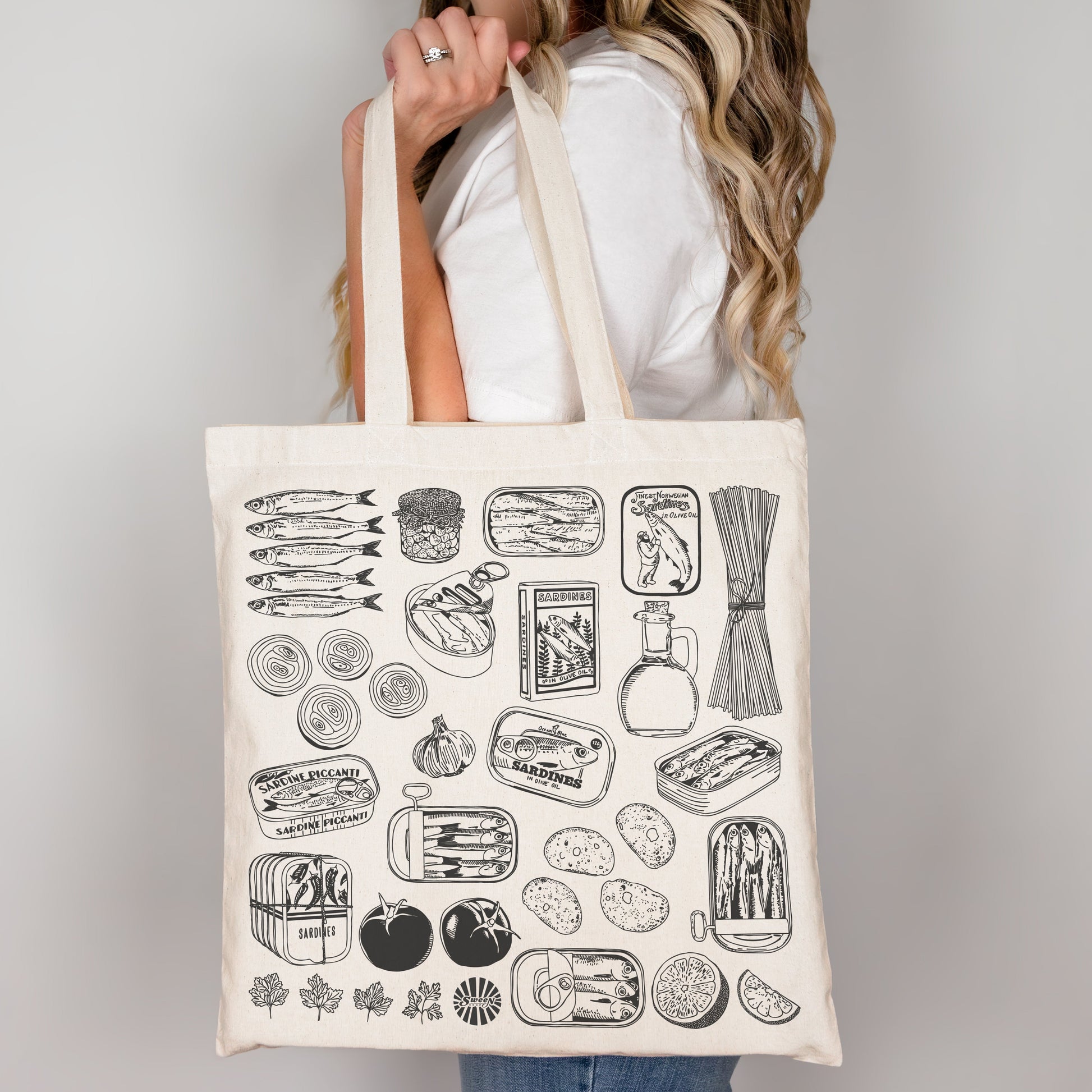 Person holding a tote bag with black sardine pasta canned fish tomato illustrations on a plain background