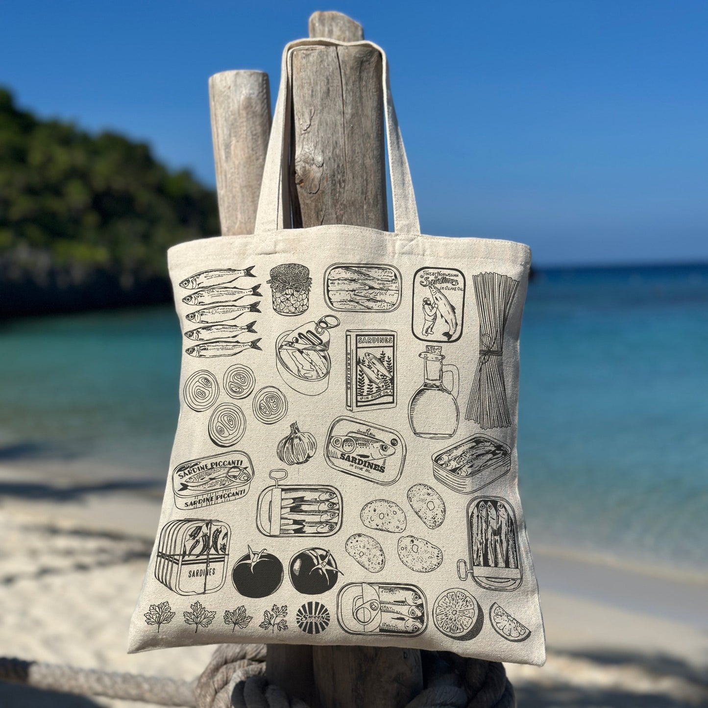 Tote bag with black sardine canned fish past tomato pattern hanging on a wooden post by the beach