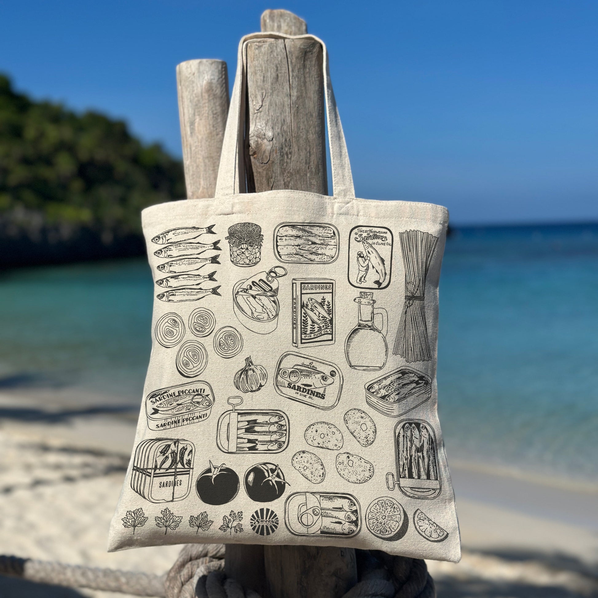 Tote bag with black sardine canned fish past tomato pattern hanging on a wooden post by the beach