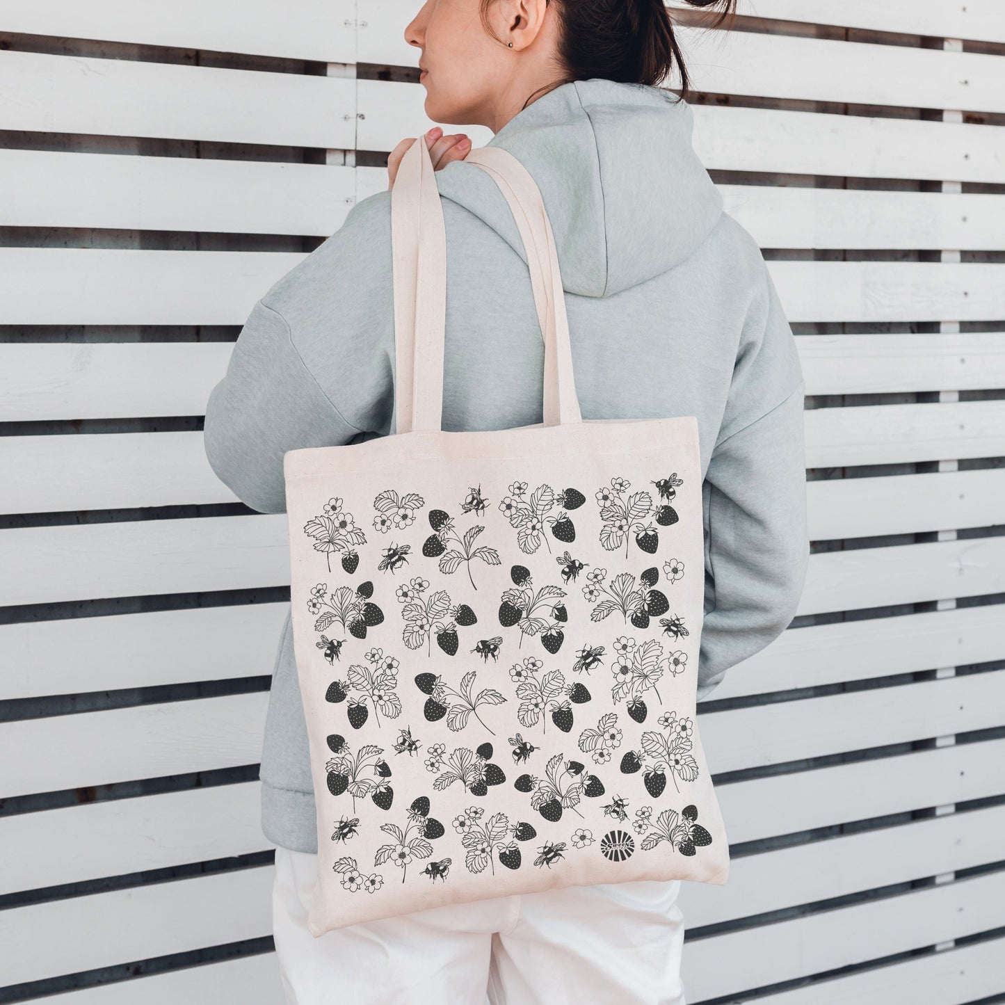 Person holding a tote bag with a floral strawberry bumble bee pattern against a white slatted wall.