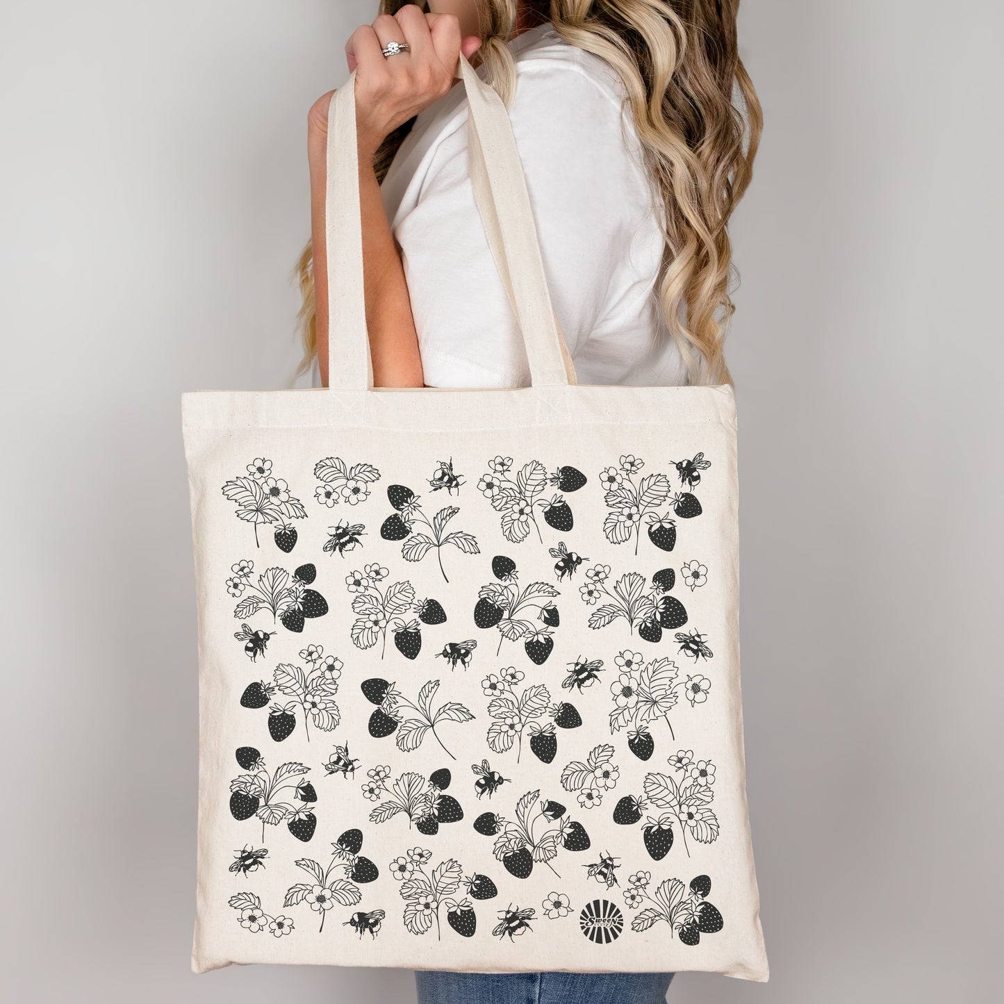 Person holding a tote bag with black floral strawberry bumble bee pattern on a plain background