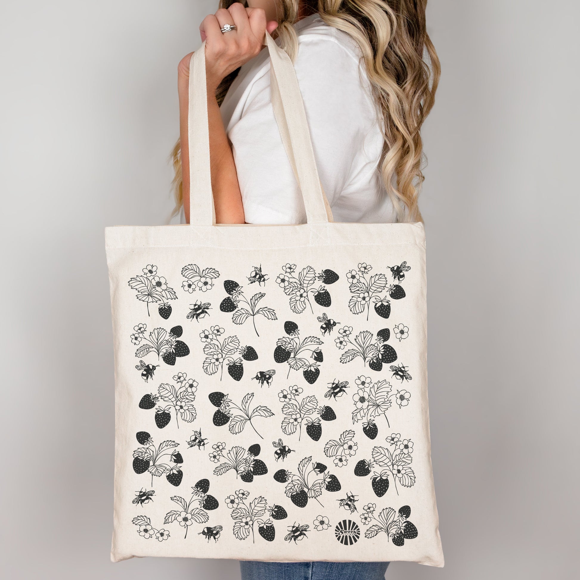 Person holding a tote bag with black floral strawberry bumble bee pattern on a plain background