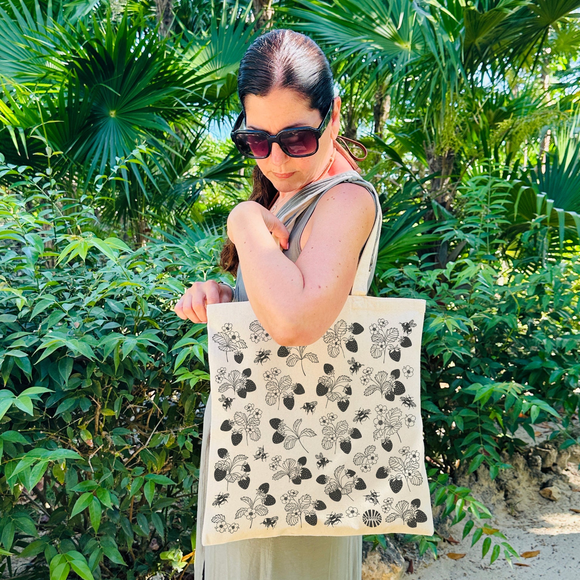 Woman holding a tote bag with a floral strawberry bumble bee pattern in a tropical beach setting