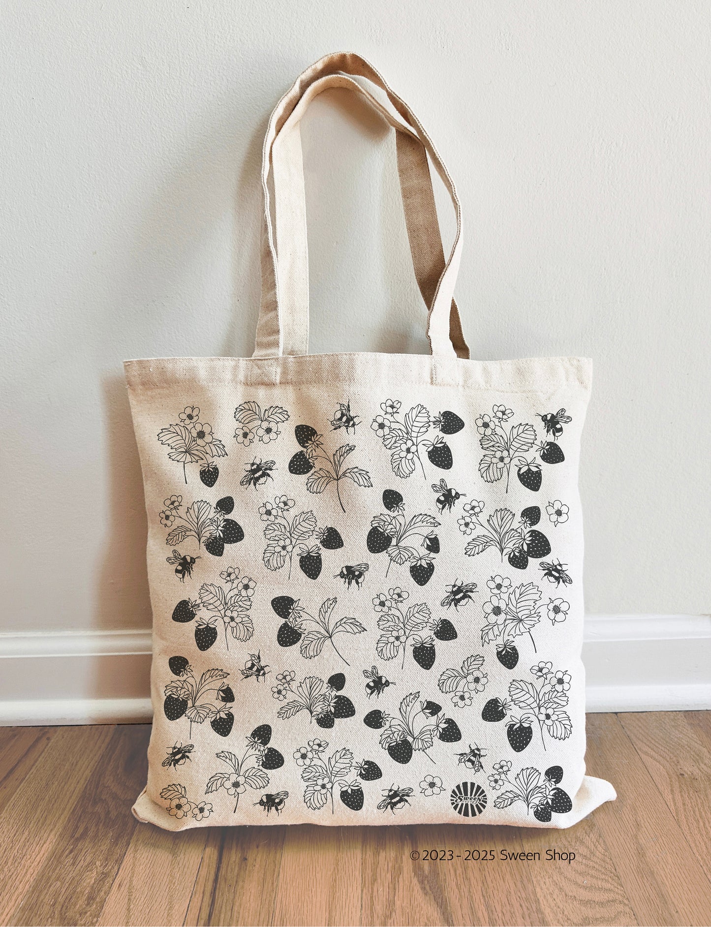 Natural cotton canvas tote bag with hand drawn black floral strawberry and bumble bee pattern on a wooden floor against a white wall.