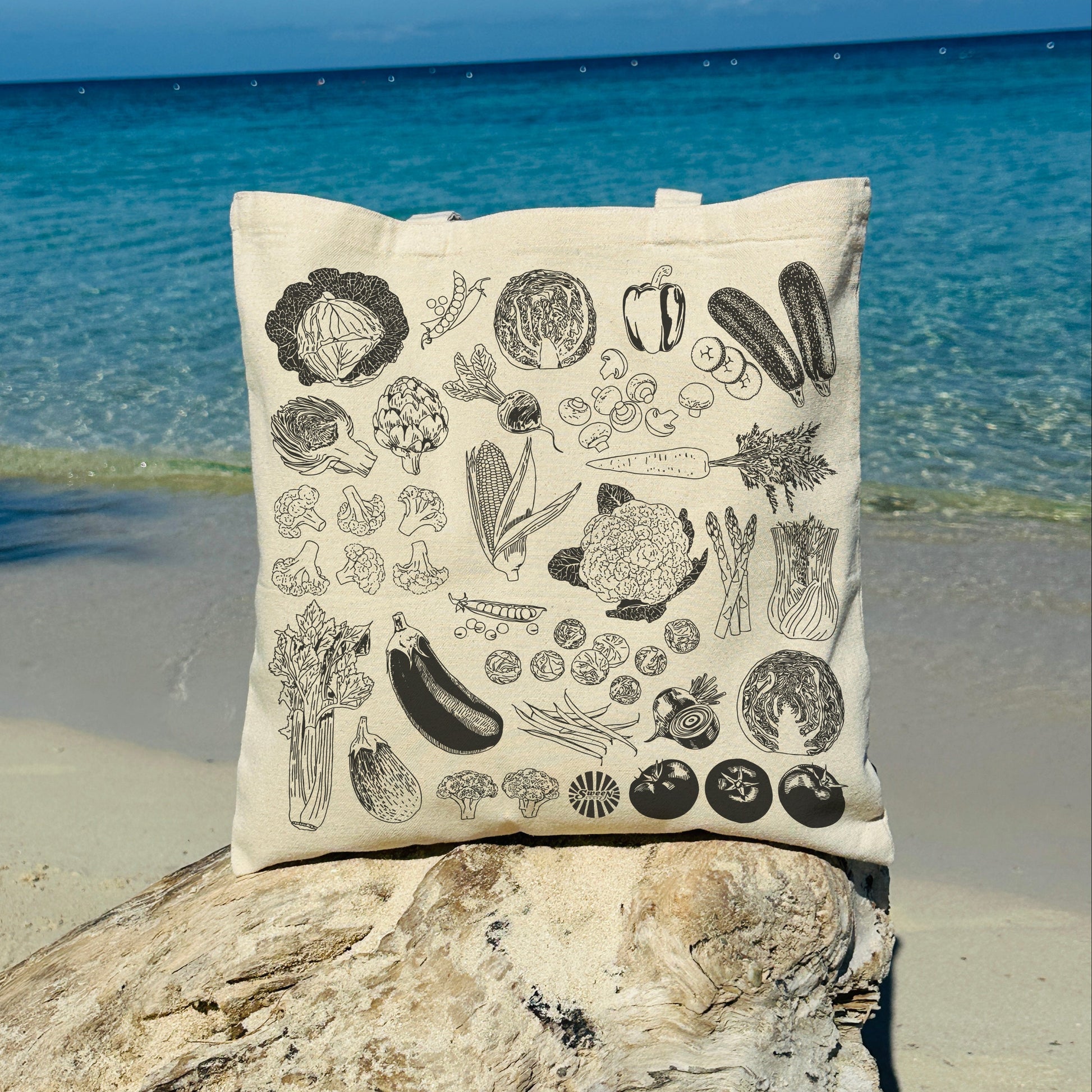 Natural cotton canvas tote bag with black outline vegetable illustrations on a stone by the ocean