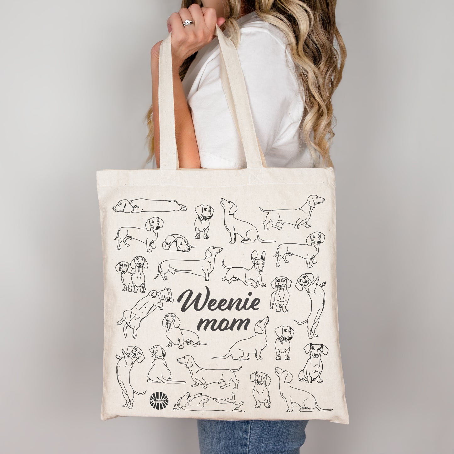 Person holding a tote bag with black line drawings of dachshund dogs and 'Weenie Mom' text on a plain background