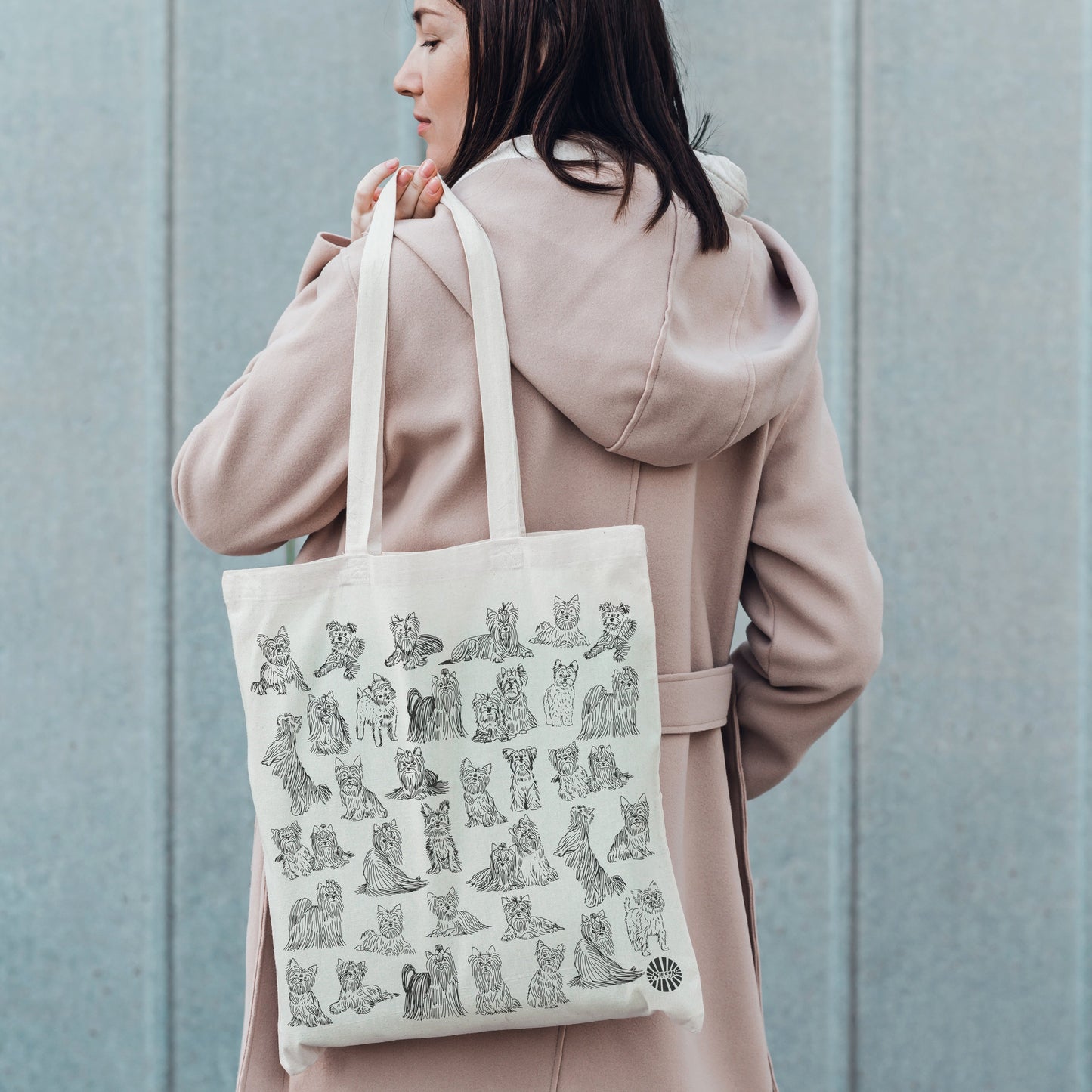 Person wearing a beige coat holding a tote bag with a printed pet yorshire terrier dog illustration against a blue wall.
